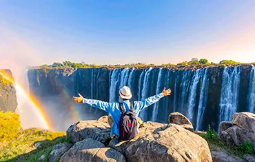 A person with a backpack raising their arms towards Victoria Falls with a rainbow in view.